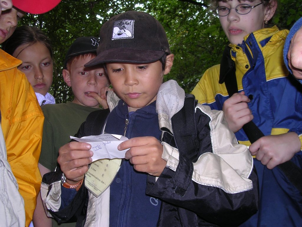 Kindergruppe im Wald schaut gemeinsam auf einen Zettel bei einer Naturaktion