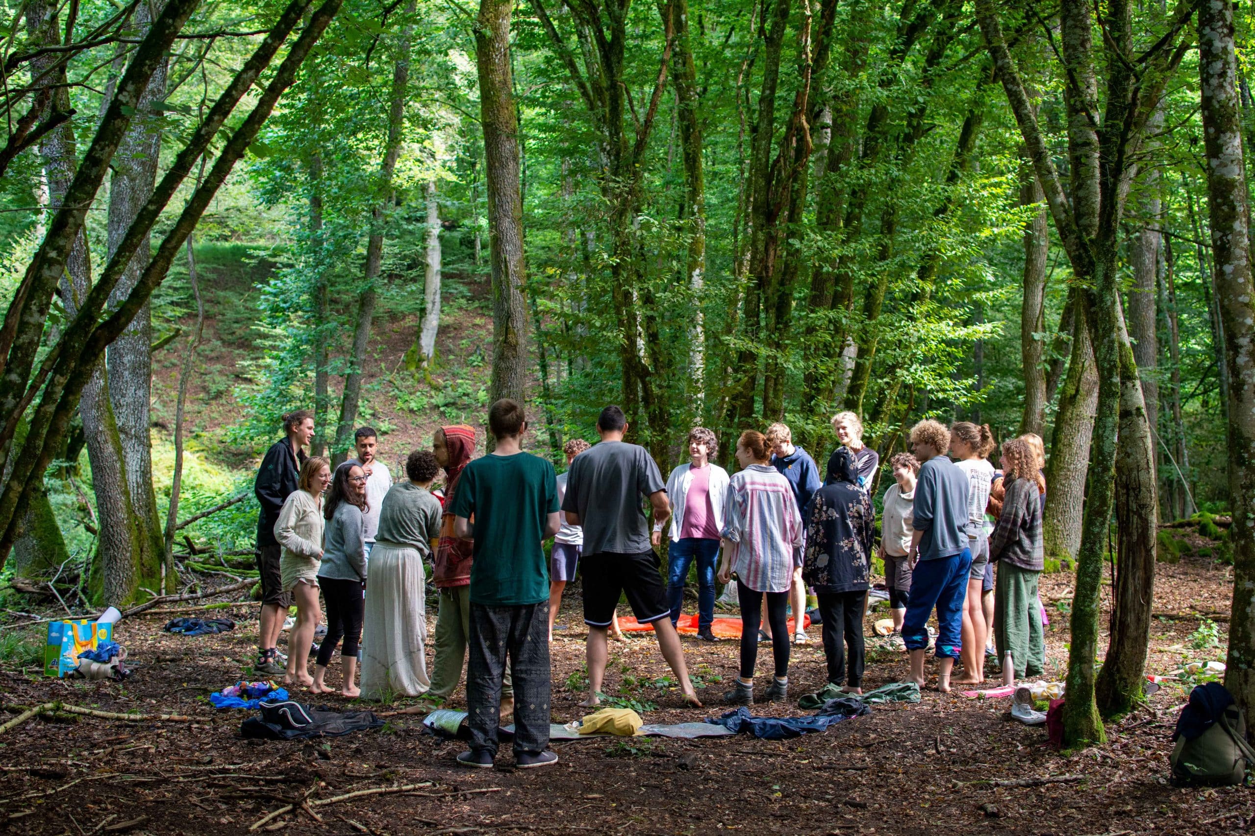 Gruppe junger Menschen steht barfuß oder locker bekleidet im Kreis auf einer Waldlichtung – Gemeinschaft und Naturverbundenheit