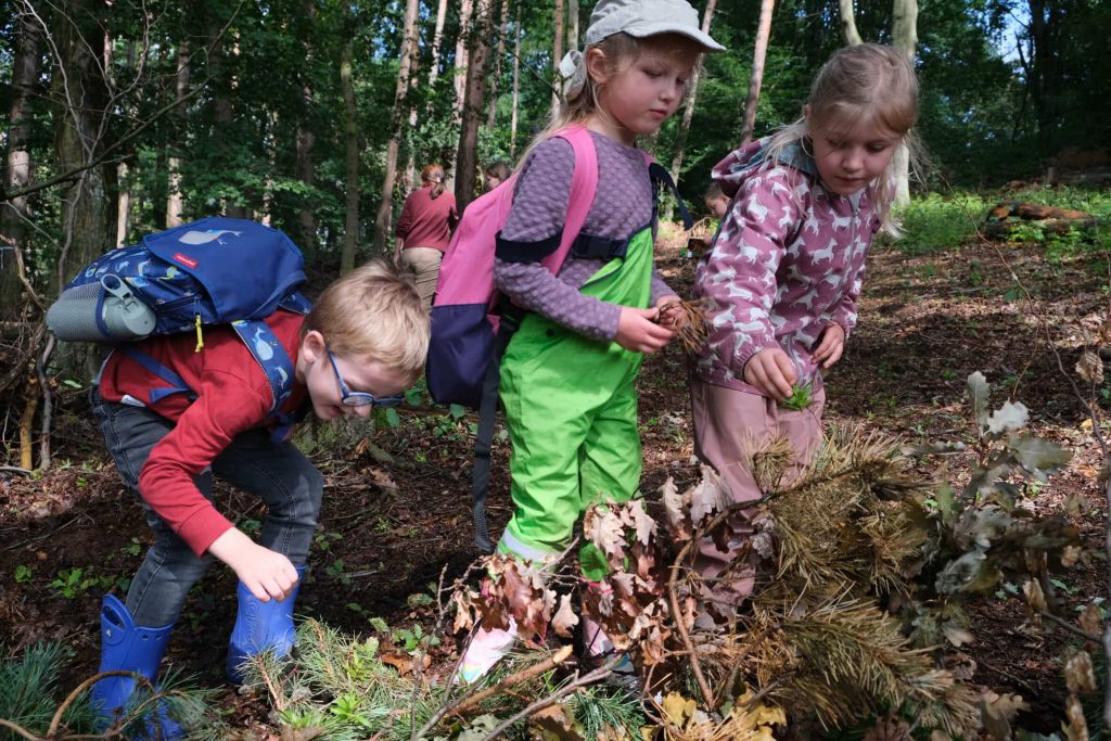 Drei Kinder mit Rucksäcken und wetterfester Kleidung sammeln Blätter und Zweige im Wald – forschend und aufmerksam im Unterholz