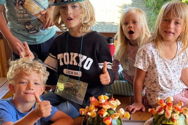 Fünf Kinder sitzen lachend um einen Tisch mit bunten Obstspießen, die in Wassermelonen gesteckt sind – gesunde, kreative Snackidee