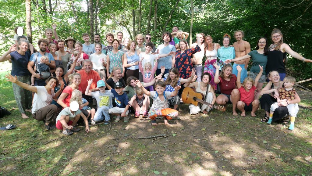 Gruppenfoto mit vielen lachenden Menschen aller Altersgruppen in einem lichten Wald &ndash; Teilnehmende des Utopiecamps winken fr&ouml;hlich in die Kamera